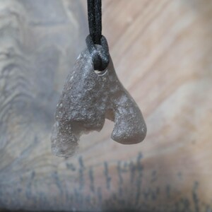 May include: A gray, organic-shaped pendant with a textured surface, suspended from a black cord. The pendant has a unique, abstract form, possibly carved from stone or a similar material. The background is a neutral, layered pattern.