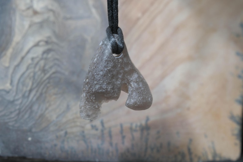 May include: A gray, organic-shaped pendant with a textured surface, suspended from a black cord. The pendant has a unique, abstract form, possibly carved from stone or a similar material. The background is a neutral, layered pattern.