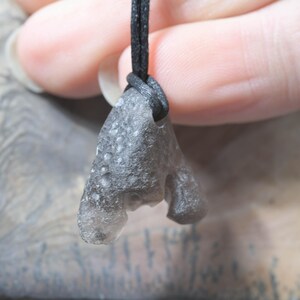 May include: A gray, irregularly shaped pendant made of a textured material, suspended from a black cord. The pendant has a unique, organic form with a rough surface. The cord is knotted at the top, securing the pendant.