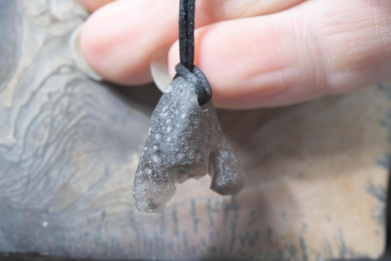 May include: A gray, irregularly shaped pendant made of a textured material, suspended from a black cord. The pendant has a unique, organic form with a rough surface. The cord is knotted at the top, securing the pendant.
