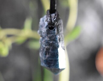 May include: A blue and gray crystal pendant hangs from a black cord. The crystal has a rough, natural texture and a faceted, polished surface that reflects light. The background is blurred, with green and gray tones.