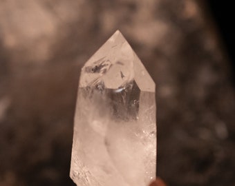 May include: A clear quartz crystal point with a natural, rough base. The crystal is sitting on a gray, textured surface.