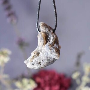 Puede incluir: Un colgante con forma de media luna hecho de una piedra gris clara, rugosa y texturizada. El colgante est&aacute; colgado de un cord&oacute;n negro.