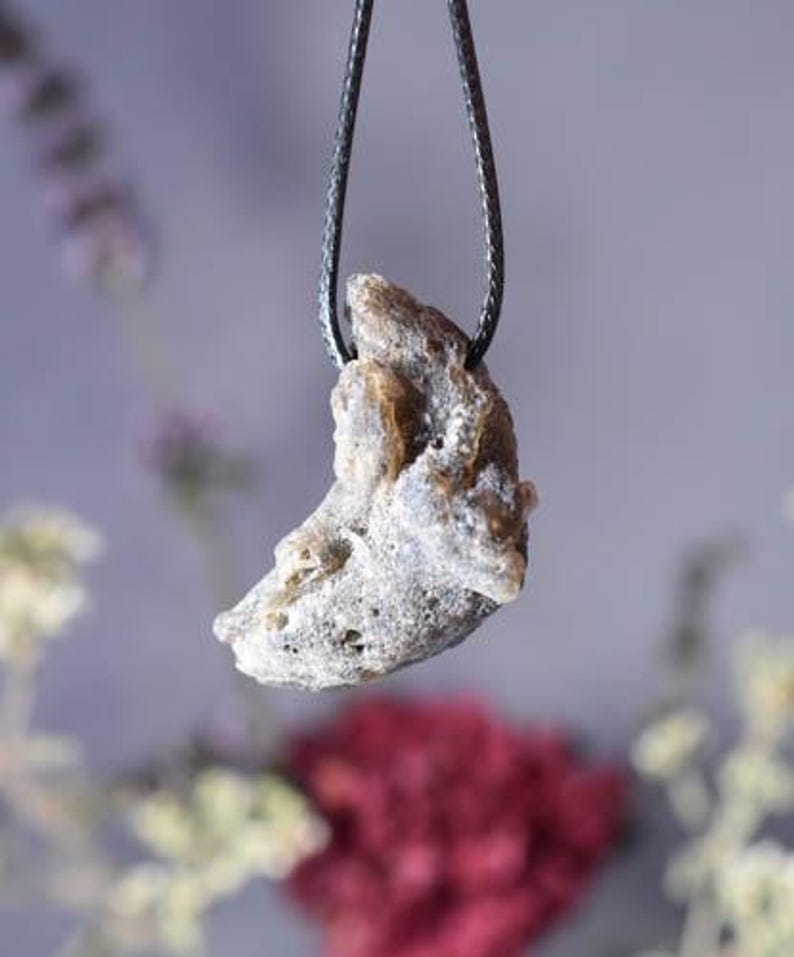 Puede incluir: Un colgante con forma de media luna hecho de una piedra gris clara, rugosa y texturizada. El colgante est&aacute; colgado de un cord&oacute;n negro.