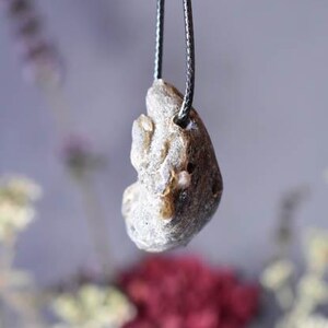 Puede incluir: Un colgante de piedra gris con una forma org&aacute;nica rugosa que cuelga de un cord&oacute;n negro. La piedra tiene un color gris claro y una superficie texturizada.