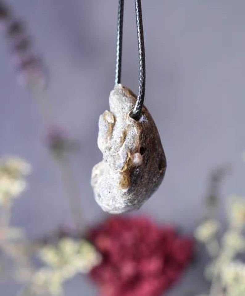 Puede incluir: Un colgante de piedra gris con una forma org&aacute;nica rugosa que cuelga de un cord&oacute;n negro. La piedra tiene un color gris claro y una superficie texturizada.