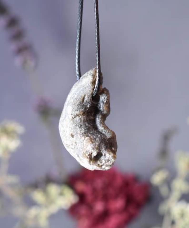 Puede incluir: Un colgante de piedra gris con una forma org&aacute;nica rugosa que cuelga de un cord&oacute;n negro. El colgante tiene un dise&ntilde;o &uacute;nico, asim&eacute;trico y una superficie texturizada.