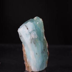 Op de afbeelding: Een groot, ruw gezaagd stuk blauwgroen aquamarijn kristal met witte strepen en bruine insluitsels.
