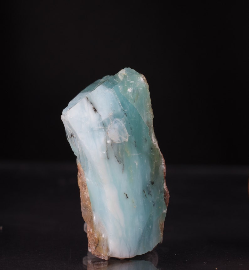 Op de afbeelding: Een groot, ruw gezaagd stuk blauwgroen aquamarijn kristal met witte strepen en bruine insluitsels.
