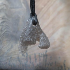 May include: A gray, irregularly shaped pendant made of a stone-like material, suspended from a black cord. The pendant has a unique, organic form with a hole for the cord. The background is a blurred mix of light and dark tones.