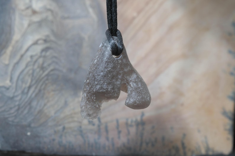 May include: A gray, irregularly shaped pendant made of a stone-like material, suspended from a black cord. The pendant has a unique, organic form with a hole for the cord. The background is a blurred mix of light and dark tones.