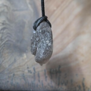 May include: A gray, textured stone pendant suspended from a black cord. The stone has a rough, natural appearance, with a slightly elongated shape. The cord is knotted around the stone, creating a secure attachment. The background is a blurred, neutral tone.