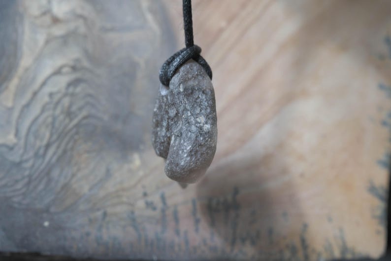May include: A gray, textured stone pendant suspended from a black cord. The stone has a rough, natural appearance, with a slightly elongated shape. The cord is knotted around the stone, creating a secure attachment. The background is a blurred, neutral tone.