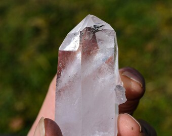 Puede incluir: Un cristal de cuarzo transparente con una punta puntiaguda y una base plana. El cristal se sostiene en una mano, y el fondo está desenfocado.