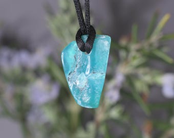 May include: A turquoise gemstone pendant with a black cord. The pendant is an irregular shape, with a polished, semi-transparent surface. The stone has a mix of light and dark blue hues with some gray inclusions.
