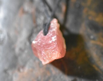 May include: A rough-cut, pink gemstone pendant suspended from a black cord. The stone has a textured surface and a natural, organic shape. The background is a blurred, mottled gray and brown.