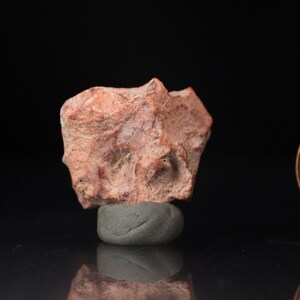 May include: A small, irregularly shaped, reddish-brown rock with a rough texture. The rock is resting on a small, gray, rounded base against a black background. The rock has a few darker red streaks.