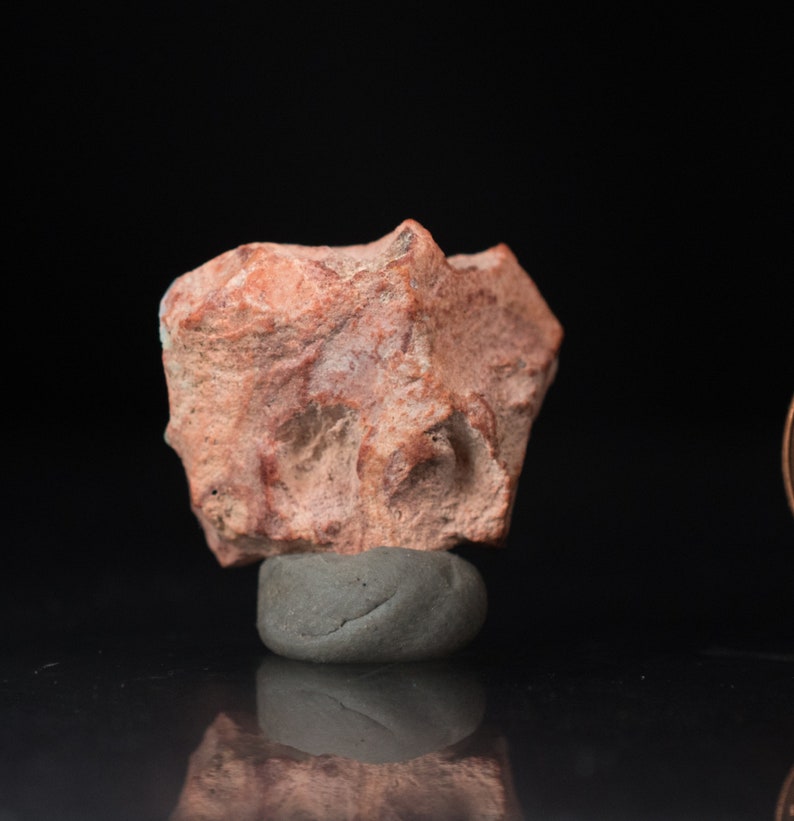 May include: A small, irregularly shaped, reddish-brown rock with a rough texture. The rock is resting on a small, gray, rounded base against a black background. The rock has a few darker red streaks.