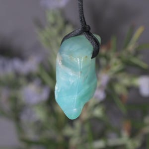 Peut inclure: Pendentif en pierre pr&eacute;cieuse bleu-vert clair, poli. Il est suspendu &agrave; un cordon noir. La pierre a une forme arrondie avec des veines blanches. L'arri&egrave;re-plan est flou, avec des touches de vert et de violet.