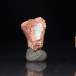 May include: A rough, reddish-brown stone with a large, white, opal-like inclusion. The stone is mounted on a small, gray, rounded rock, set against a black background. The opal inclusion has a pearly, iridescent quality.
