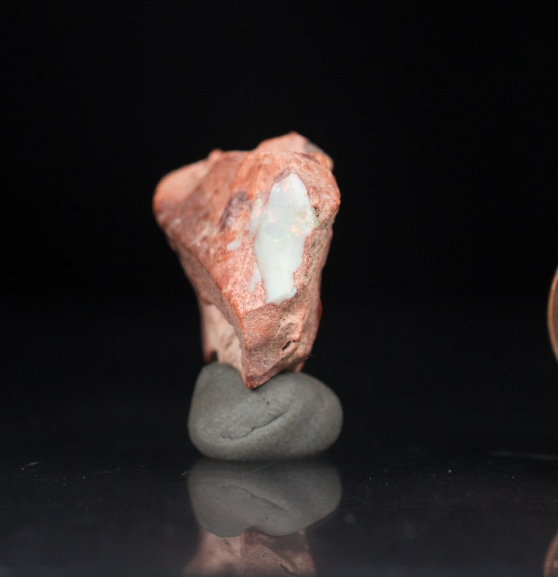 May include: A rough, reddish-brown stone with a large, white, opal-like inclusion. The stone is mounted on a small, gray, rounded rock, set against a black background. The opal inclusion has a pearly, iridescent quality.
