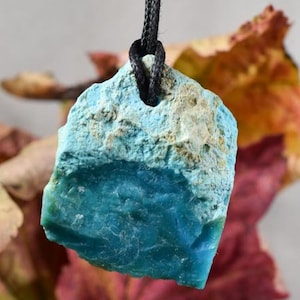 Może przedstawiać: Wisiorek z naturalnego kamienia w kolorze turkusowym i beżowym na czarnym sznurku. Wisiorek ma nieregularny kształt z teksturowaną powierzchnią. Kamień ma ciemniejszy turkusowy odcień na dole. Wisiorek zawieszony na rozmytym tle.
