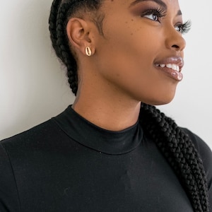 Peut inclure: Une femme à la peau brune foncée et aux cheveux noirs tressés porte un haut noir à col montant. Elle regarde vers la droite et sourit. Elle porte des boucles d'oreilles en forme de coquille dorées.