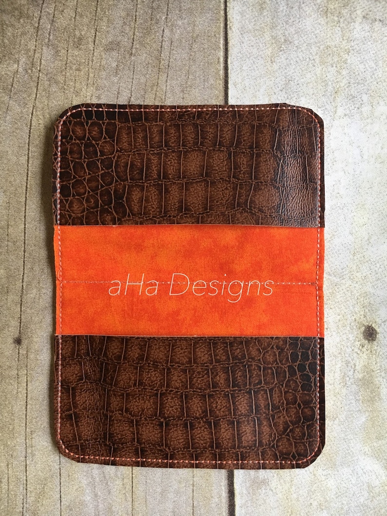 May include: A brown leather passport holder with a crocodile embossed pattern and a bright orange suede insert. The text "aHa Designs" is printed in white on the orange suede.