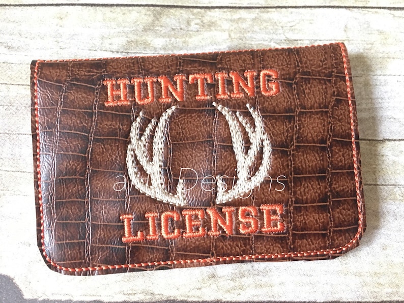 May include: Brown faux leather hunting license holder with an embroidered orange "HUNTING" and "LICENSE" with a white embroidered antler design in the center.