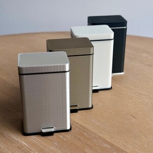 May include: Four miniature trash cans in different colors: silver, gold, white, and black. The cans are lined up on a wooden surface.
