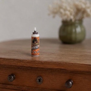 May include: A miniature replica of a Gorilla Wood Glue bottle featuring a white cap, an orange label with a gorilla graphic, and the text Gorilla Wood Glue. The small cylindrical container stands upright with a pointed nozzle tip at the top.