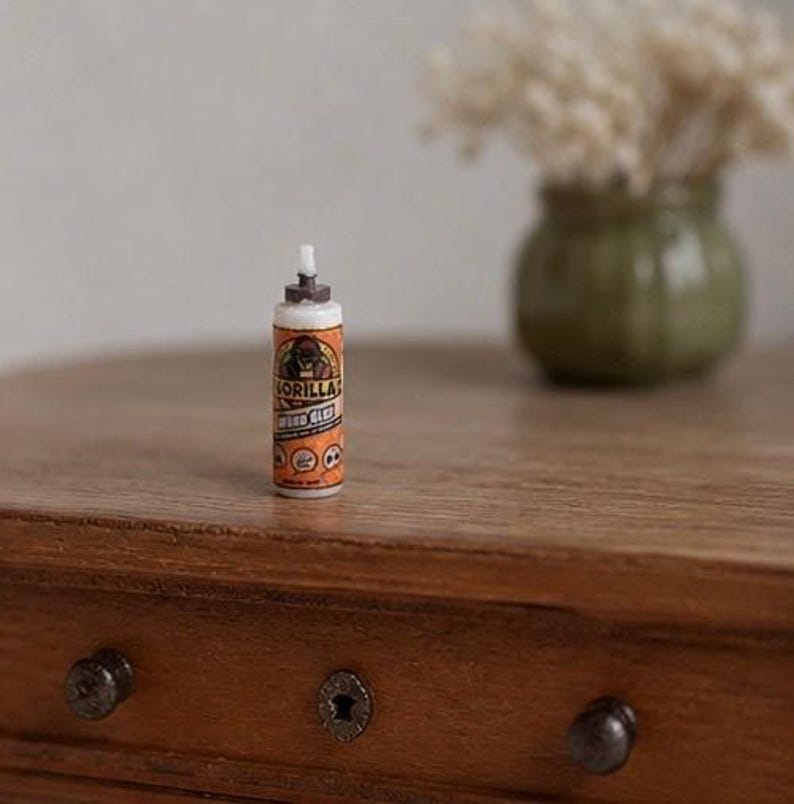 May include: A miniature replica of a Gorilla Wood Glue bottle featuring a white cap, an orange label with a gorilla graphic, and the text Gorilla Wood Glue. The small cylindrical container stands upright with a pointed nozzle tip at the top.