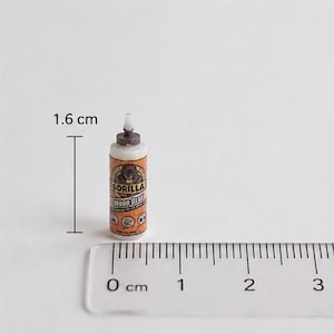 May include: A miniature bottle of Gorilla Wood Glue with an orange and white label featuring a gorilla logo. The bottle has a brown cap and a white nozzle. A clear plastic ruler is positioned at the base, and a measurement of 1.6 cm is indicated.