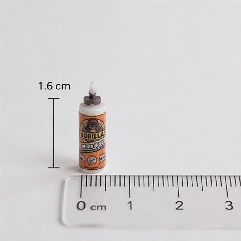 May include: A miniature bottle of Gorilla Wood Glue with an orange and white label featuring a gorilla logo. The bottle has a brown cap and a white nozzle. A clear plastic ruler is positioned at the base, and a measurement of 1.6 cm is indicated.