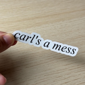 May include: A white sticker with the text "carl's a mess" in a black, elegant font. The sticker is held by a hand against a light brown wooden surface. The sticker is a humorous statement.