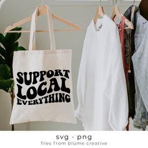 Op de afbeelding: Een crèmekleurige draagtas met de zwarte tekst "SUPPORT LOCAL EVERYTHING". De tas hangt aan een kledingrek met andere kledingstukken. De tas is gemaakt van een natuurlijke stof.