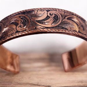 May include: A copper bracelet with an intricate engraved floral design. The bracelet is open and has a smooth, polished finish.