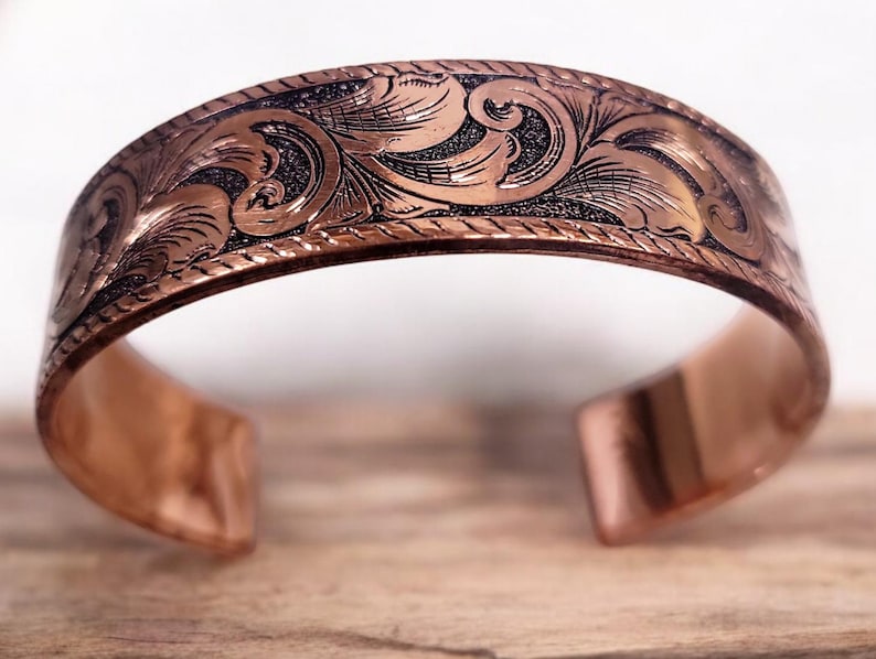 May include: A copper bracelet with an intricate engraved floral design. The bracelet is open and has a smooth, polished finish.