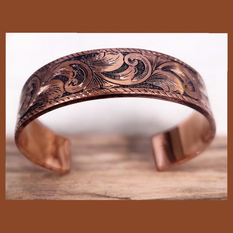 May include: A copper bracelet with an intricate engraved floral design. The bracelet is open and has a smooth, polished finish.