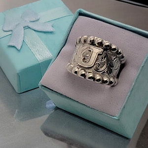 May include: A silver ring with a beaded edge and an engraved floral design featuring the letter "J" in the center. The ring is displayed in a light blue gift box.
