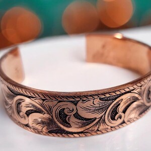 May include: A copper bracelet with an intricate floral design. The bracelet is made of a smooth, polished copper and has a slightly curved shape.