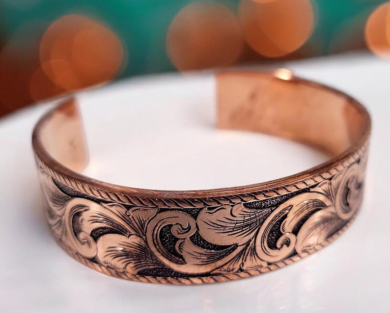May include: A copper bracelet with an intricate floral design. The bracelet is made of a smooth, polished copper and has a slightly curved shape.