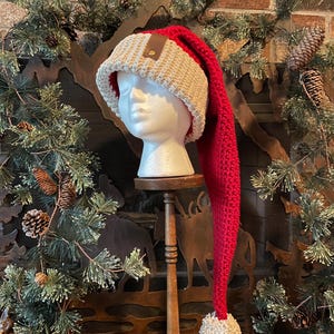 May include: A long, red crocheted Santa hat with a white pom-pom and a brown leather patch on the top. The hat is on a white mannequin head.