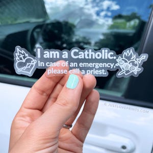 May include: A clear rectangular sticker with white text that reads, I am a Catholic. In case of an emergency, please call a priest. The sticker features a white line art illustration of the Sacred Heart on the left and the Immaculate Heart on the right.