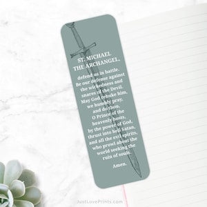 May include: A rectangular bookmark with rounded corners, featuring a prayer to St. Michael the Archangel. The text is in white on a muted teal background, with a detailed illustration of a sword. The bookmark is resting on a lined paper surface.