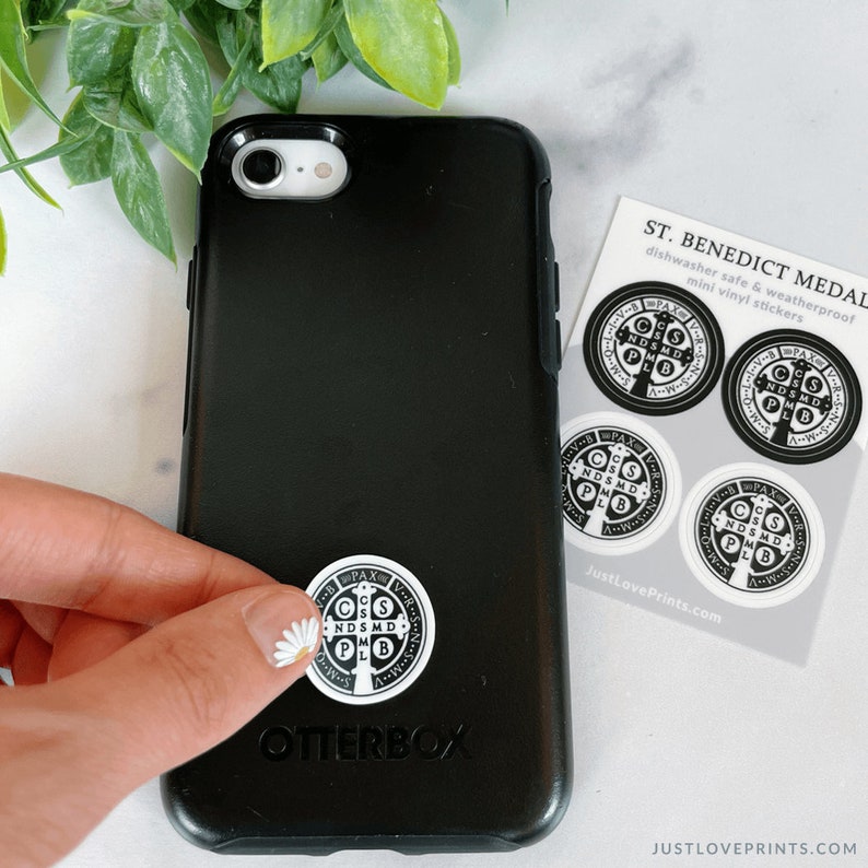 These sticker sheets contain 4 St Benedict medal stickers, in black and white. They are dishwasher safe and weatherproof mini vinyl stickers. The photo shows Lindsay placing a medal on a phone case.