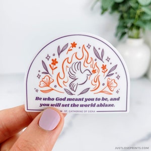 May include: A white sticker with a purple and orange illustration of a dove in flames. The sticker has the quote "Be who God meant you to be, and you will set the world ablaze." and the text "ST. CATHERINE OF SIENA".