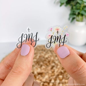 Op de afbeelding: Twee transparante stickers met de tekst "+ JMJ" in zwarte schrijfletters. E&eacute;n sticker heeft een aquarel bloemenpatroon met roze, gele en witte bloemen.