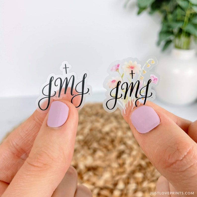 Op de afbeelding: Twee transparante stickers met de tekst "+ JMJ" in zwarte schrijfletters. E&eacute;n sticker heeft een aquarel bloemenpatroon met roze, gele en witte bloemen.