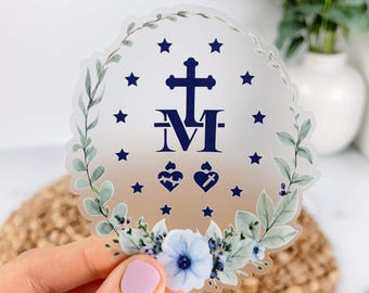 May include: A circular sticker featuring a dark blue cross and the letter M, flanked by two small hearts and surrounded by a ring of twelve dark blue stars. The design is framed by a watercolor-style wreath of green leaves and light blue flowers.